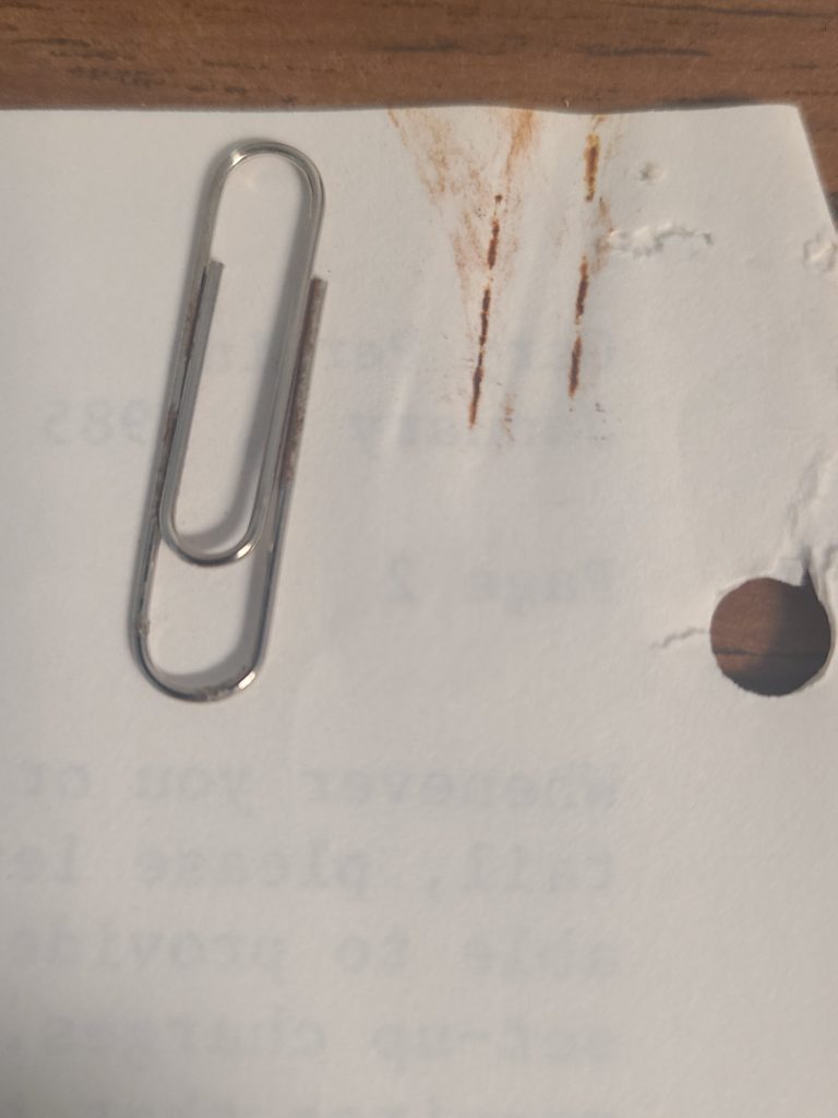A paperclip on white paper. Next to the paperclip is a red-orange imprint of it from rust damage,