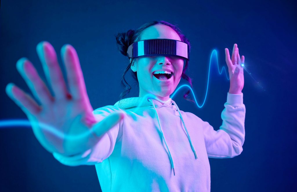 An Adobe Stock Photo in color of a woman in a VR headset smiling with her hands out. The colors have a vaporwave and neon tone.